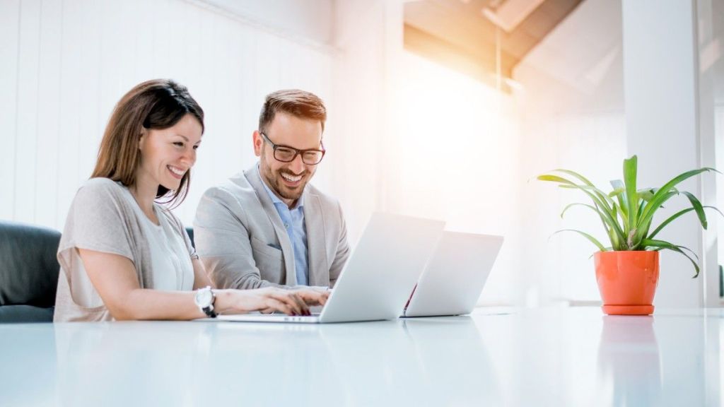 A young brunette female and a young man with glasses smile as they sit next to each other at a desk, typing on their laptops while discussing how to manage business expenses