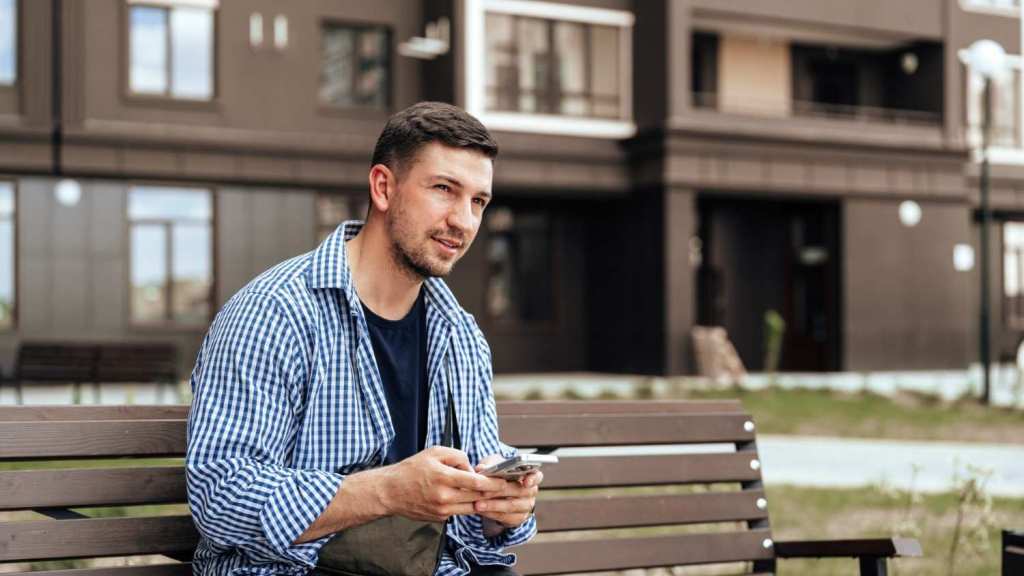 A young man in a blue check shirt looks ahead as he sits on a bench outside and reads about the Revolut Business Account on his mobile phone